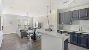 A modern kitchen with dark cabinets, a white countertop, and a dining area with a gray sofa and chairs. - Landing | Modern 1BD, Pool, Courtyard (Cary)