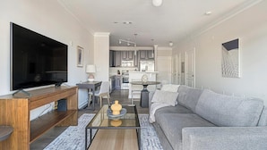 A modern living room with a flat-screen TV, a wooden coffee table, a grey sofa, and a kitchen in the background. - Landing | Modern 1BD, Pool, Courtyard (Cary)