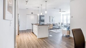 A modern kitchen with a central island, a dining area with a sofa and coffee table, and a living room with a TV and seating. - Landing | Beautiful 2BD, Gym, Pool (Tampa)