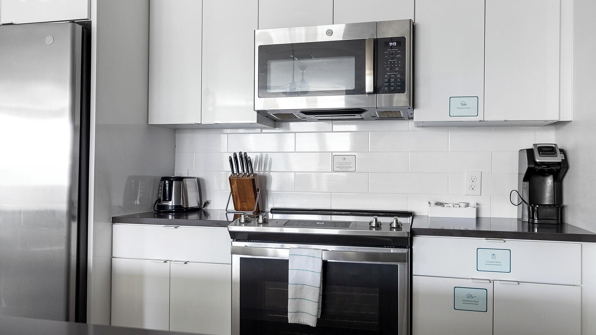 Una cocina moderna con electrodomésticos de acero inoxidable, incluyendo un microondas, horno y cafetera, con armarios blancos y salpicadero de azulejos.