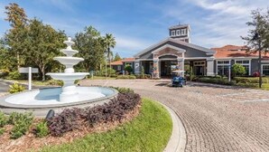 A building with a fountain in the foreground, a paved driveway, and a well-maintained lawn. - Landing | Beautiful 2BD, Courtyard, Gym (Kissimmee)