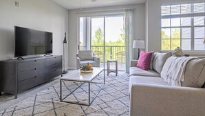 A living room with a flat-screen TV, grey sofa, coffee table, and a balcony with a chair and table. - Landing | Beautiful 2BD, Courtyard, Gym (Kissimmee)