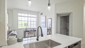 A modern kitchen with a large island, stainless steel sink, and a dishwasher. There is a dining area with a table and chairs visible through the window.