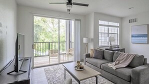 A modern living room with a grey sofa, a coffee table, and a TV on a stand. There is a balcony with a view of greenery.