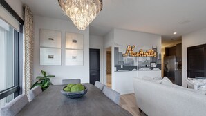 Un comedor moderno con una mesa grande, sillas tapizadas en gris y una araña. Hay cuadros en la pared, una planta y un letrero de neón.