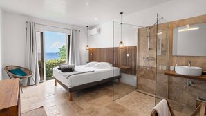 Un dormitorio moderno con cama, cabina de ducha de vidrio, mesita de noche de madera y vista al mar.