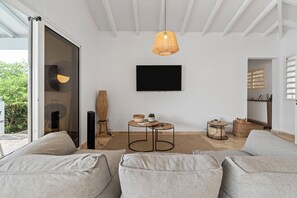 A modern living room with a sofa, a television mounted on the wall, and a hanging light fixture. - Villa La Sucrerie - Three Bedroom Villa, Sleeps 6 (Les Anses-d'Arlet)