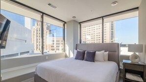 A modern hotel room with a large bed, a nightstand, and a view of city buildings through large windows. - Landing | Amazing 2BD in Fountain Heights (Birmingham)