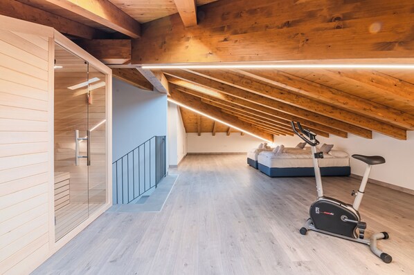 Un dormitorio tipo loft moderno con techo inclinado, vigas de madera, una puerta de vidrio que da a un balcón y una bicicleta estática.