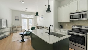 A modern kitchen with white cabinets, a large island with a sink, a microwave, and a stove. There is a dining area with a table and chairs, and a window with blinds.