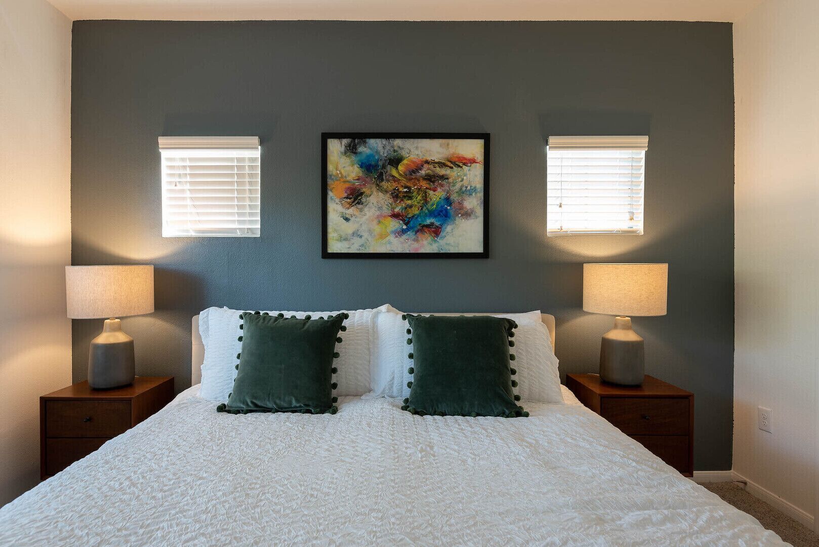 A bedroom with a large bed, two bedside lamps, a colorful abstract painting, and two windows with blinds.