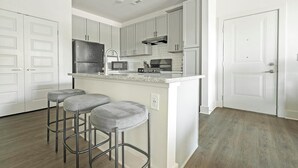 A kitchen with white cabinets, a stainless steel refrigerator, a microwave, a stove, and a sink. There are three grey bar stools. - Landing | Incredible 1BD, Clubhouse, Gym (Charlotte)