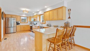 Una cocina moderna con armarios de madera, un refrigerador de acero inoxidable, una isla central con taburetes y un ventanal que proporciona luz natural.