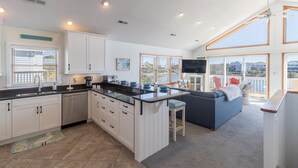 Una cocina moderna con armarios blancos, encimera negra y una isla grande. Hay una zona de comedor con un sofá azul y televisor. La habitación tiene ventanas amplias y un balcón con vistas a la ciudad.