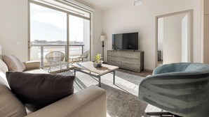A modern living room with a sofa, a coffee table, and a TV on a wooden cabinet. There is a balcony with a view of the city. - Landing | Stylish 2BD, Gym, Pool (Las Vegas)