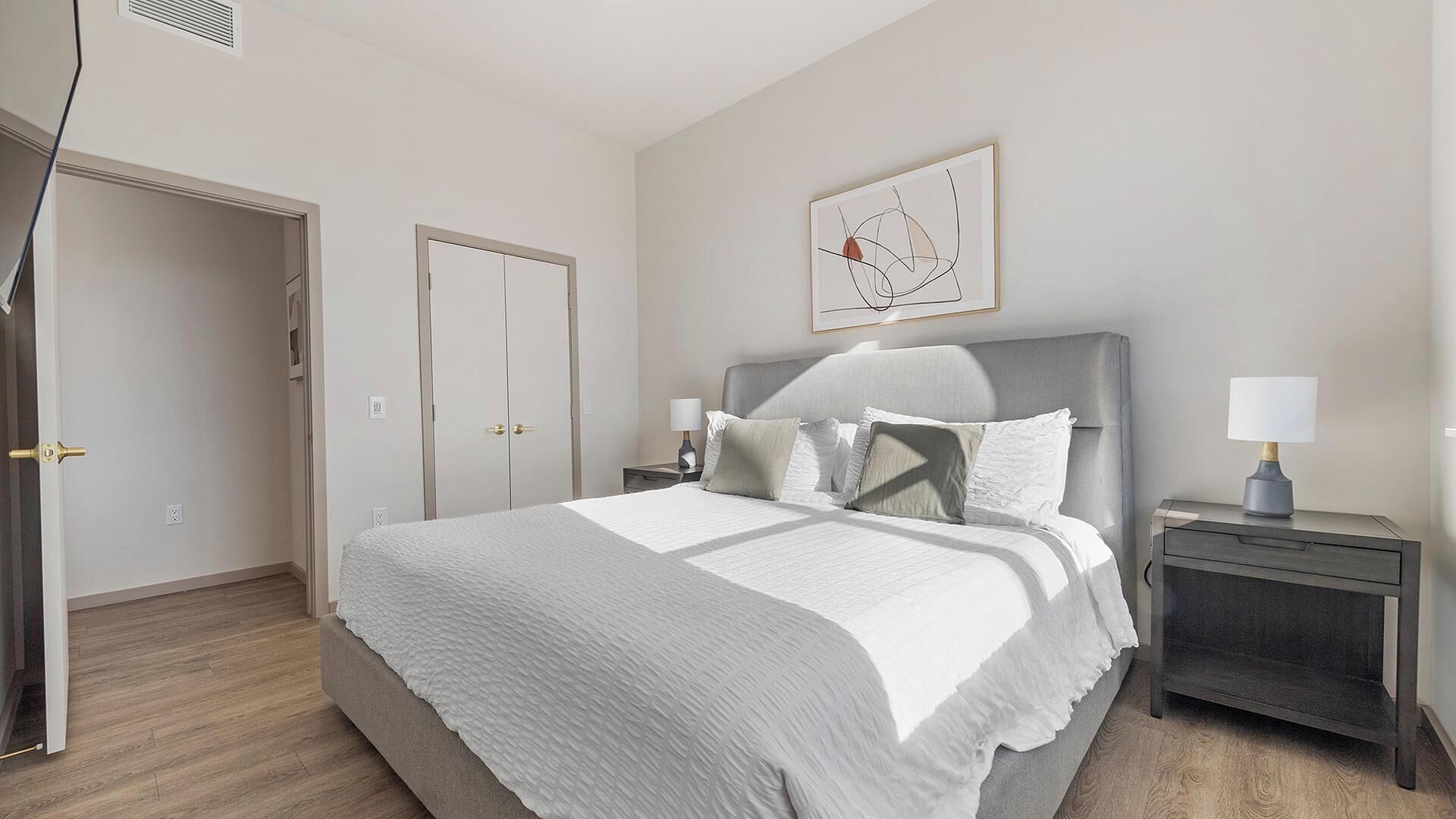 A bedroom with a bed, bedside tables, a nightstand, a door, and a wall-mounted artwork.