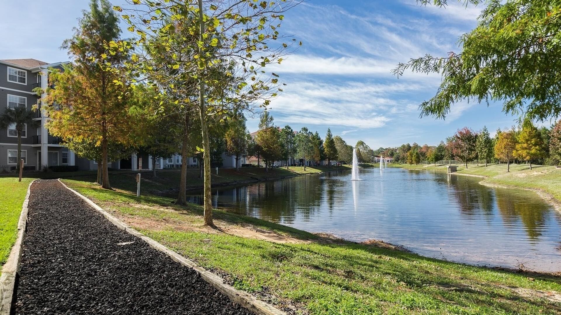 A serene riverside view with a walking path, trees, and a building complex in the background.