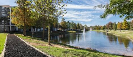 A serene riverside view with a walking path, trees, and a building complex in the background.