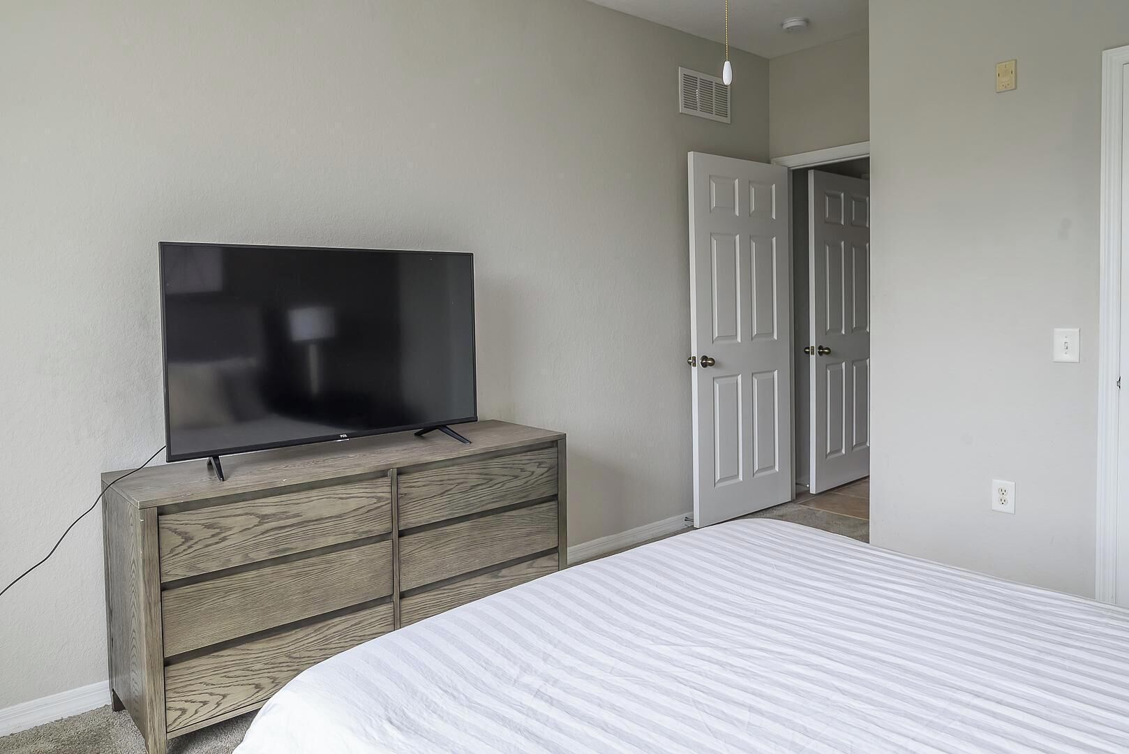 A bedroom with a bed, a wooden dresser with a TV, and a door leading to another room.