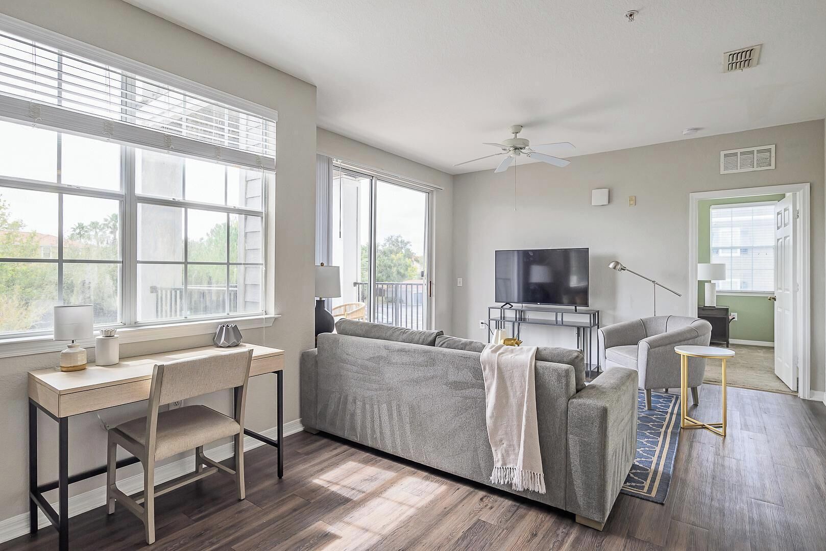 A modern living room with a grey sofa, a TV on a stand, a desk with a chair, and large windows.