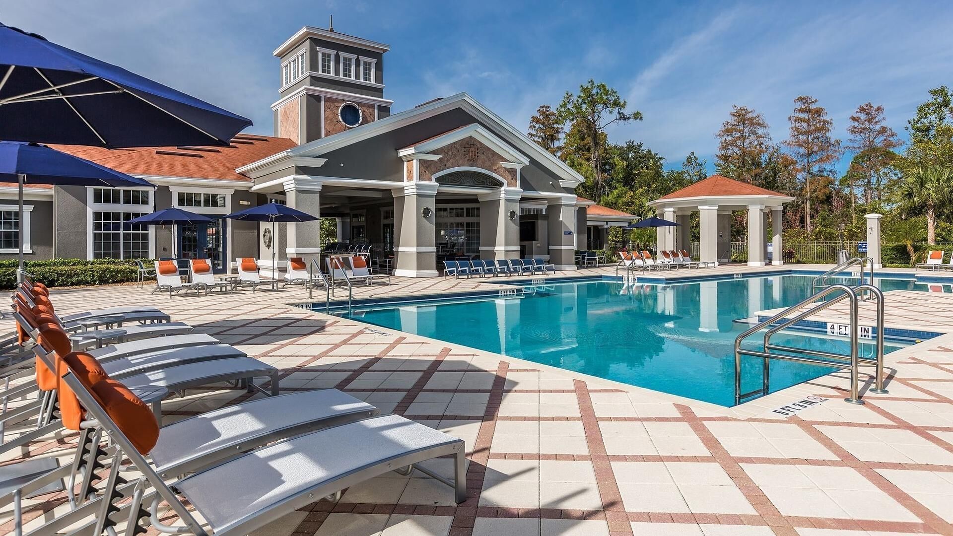 Un área de piscina con sillones de descanso y sombrillas, un edificio con una torre y un cielo azul despejado.
