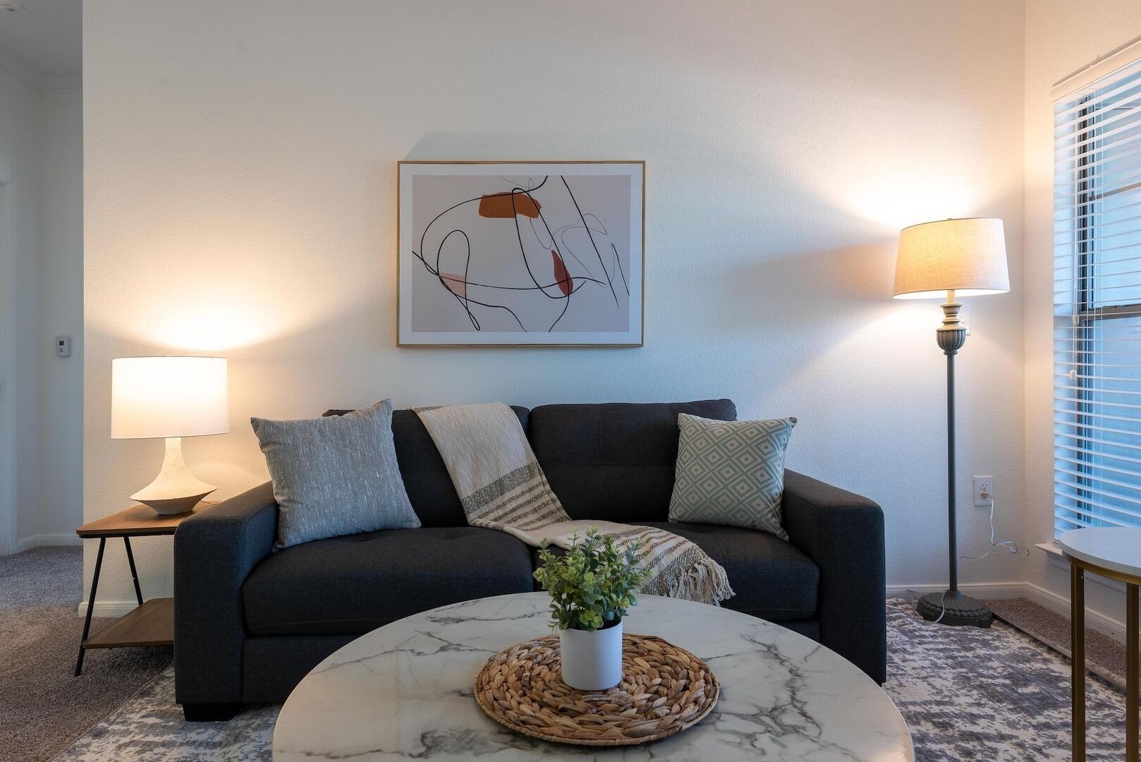 A living room with a dark sofa, a round marble coffee table, a potted plant, and a framed abstract artwork on the wall.