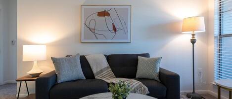 A living room with a dark sofa, a round marble coffee table, a potted plant, and a framed abstract artwork on the wall.