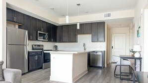 A modern kitchen with dark cabinets, stainless steel appliances, a central island, and a dining area with a table and chairs. - Landing | Amazing 2BD, Yoga Studio, Pool (Palmetto)