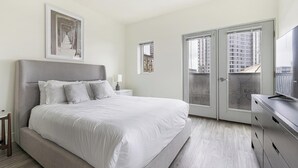 A modern bedroom with a large bed, a nightstand, a desk, and a balcony with a view of buildings. - Landing | Stylish Studio, Courtyard, Gym (Portland)