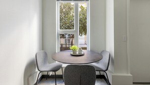 Uma área de refeições com uma mesa redonda, duas cadeiras cinzentas e uma janela com vista para vegetação e um edifício.