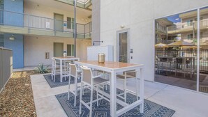 A courtyard with two white tables and chairs, a decorative urn, and a patterned rug. - Landing | Amazing 1BD, Pool, Clubhouse (Goodyear)