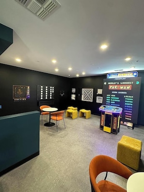 Een kamer met een Pac-Man spelcomputer, een tafel met stoelen en een muur waarop de hoogste scores staan.