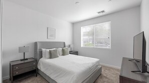 A modern bedroom with a bed, bedside tables, a TV, and a window with blinds.