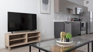 A modern living room with a flat-screen TV on a wooden stand, a glass coffee table with decorative items, and a kitchen area with stainless steel appliances. - Landing | Amazing 1BD, Pool, Clubhouse (Goodyear)