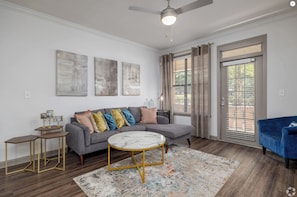 Ein modernes Wohnzimmer mit einem grauen Sofa, einem runden Couchtisch und einem blauen Sessel. An der Wand befinden sich abstrakte Gemälde und ein Deckenventilator.