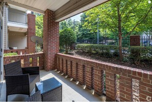 A covered outdoor seating area with wicker furniture, brick walls, and a view of a landscaped garden and street.