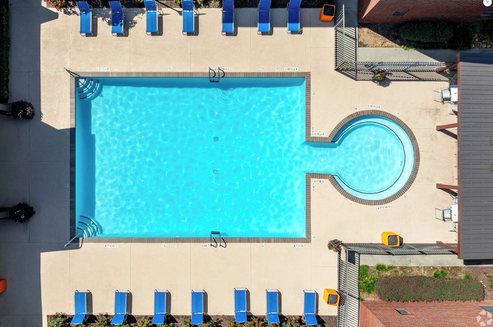 A rectangular swimming pool with a circular hot tub, surrounded by blue lounge chairs and a covered seating area.
