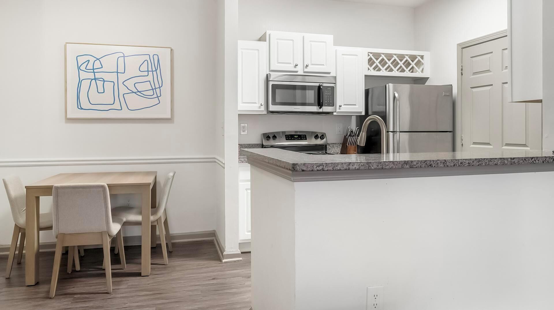 A modern kitchen with white cabinetry, a stainless steel refrigerator, a microwave, and a stove. There is a dining table with chairs and a framed abstract painting on the wall.