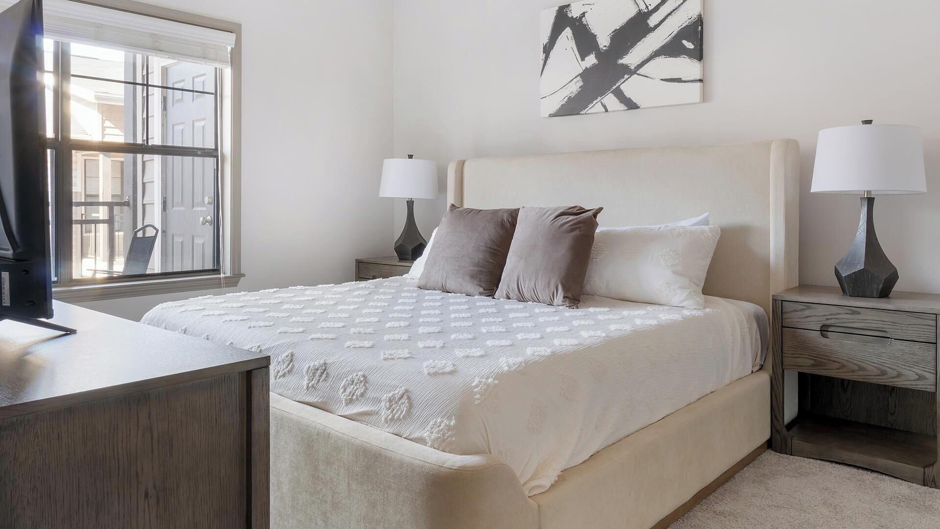 A bedroom with a large bed, two bedside tables with lamps, a window with blinds, and a wall-mounted abstract art piece.