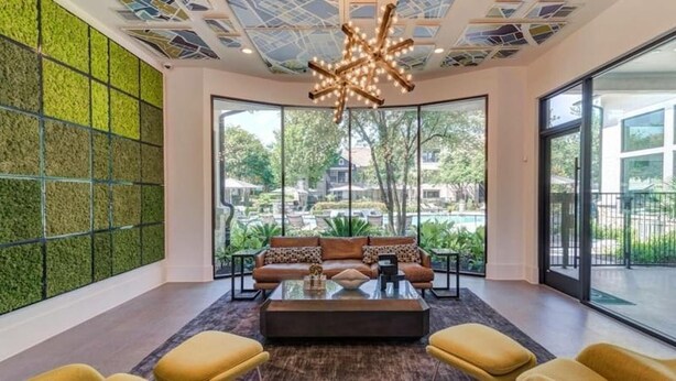 Een moderne lounge met een glazen salontafel, gele fauteuils en een bank met gekleurde kussens. Een opvallend plafondontwerp met een ster-vormige lamp is zichtbaar.