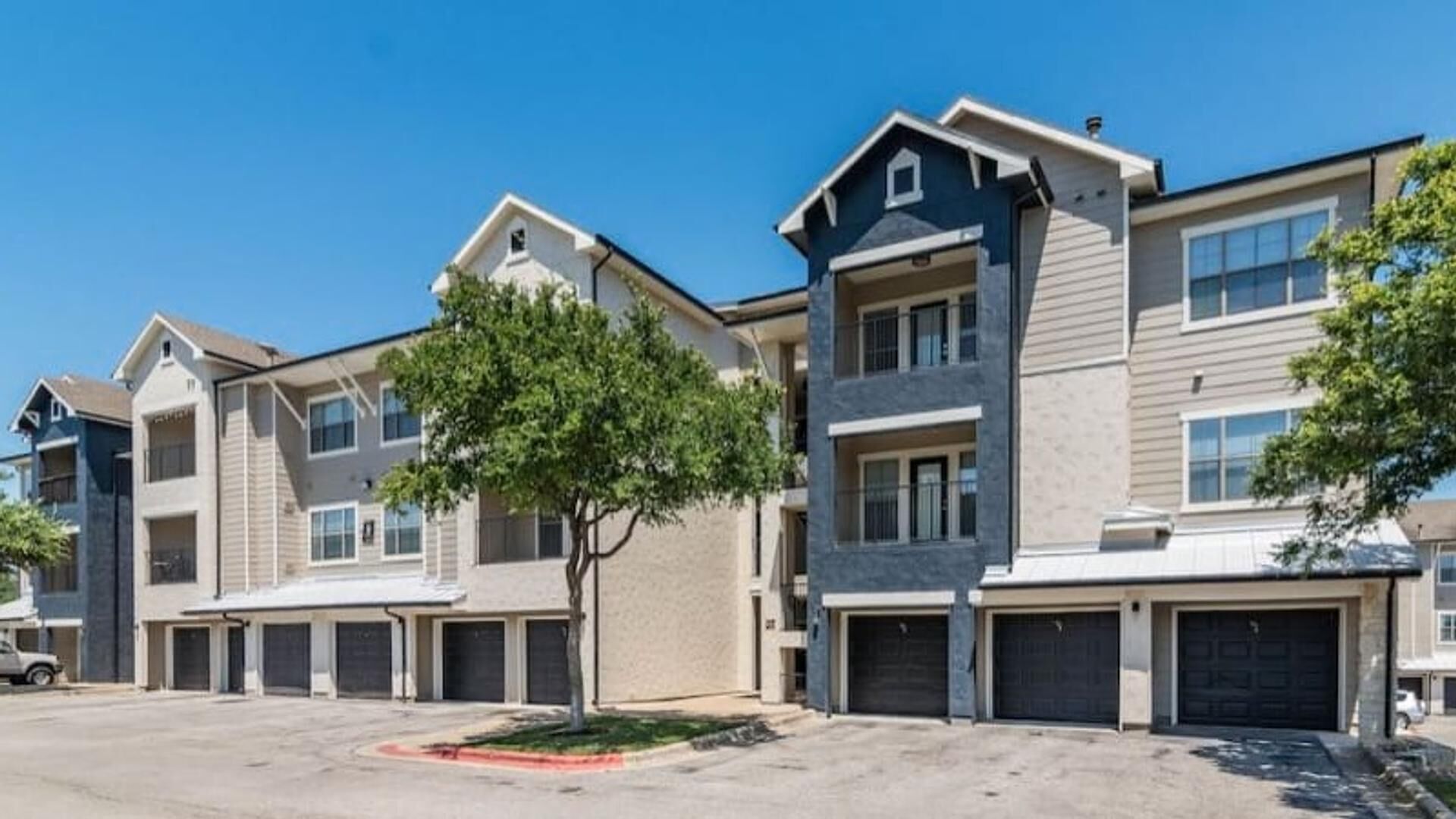 A row of modern apartment buildings with multiple garage doors, a central tree, and a clear blue sky.