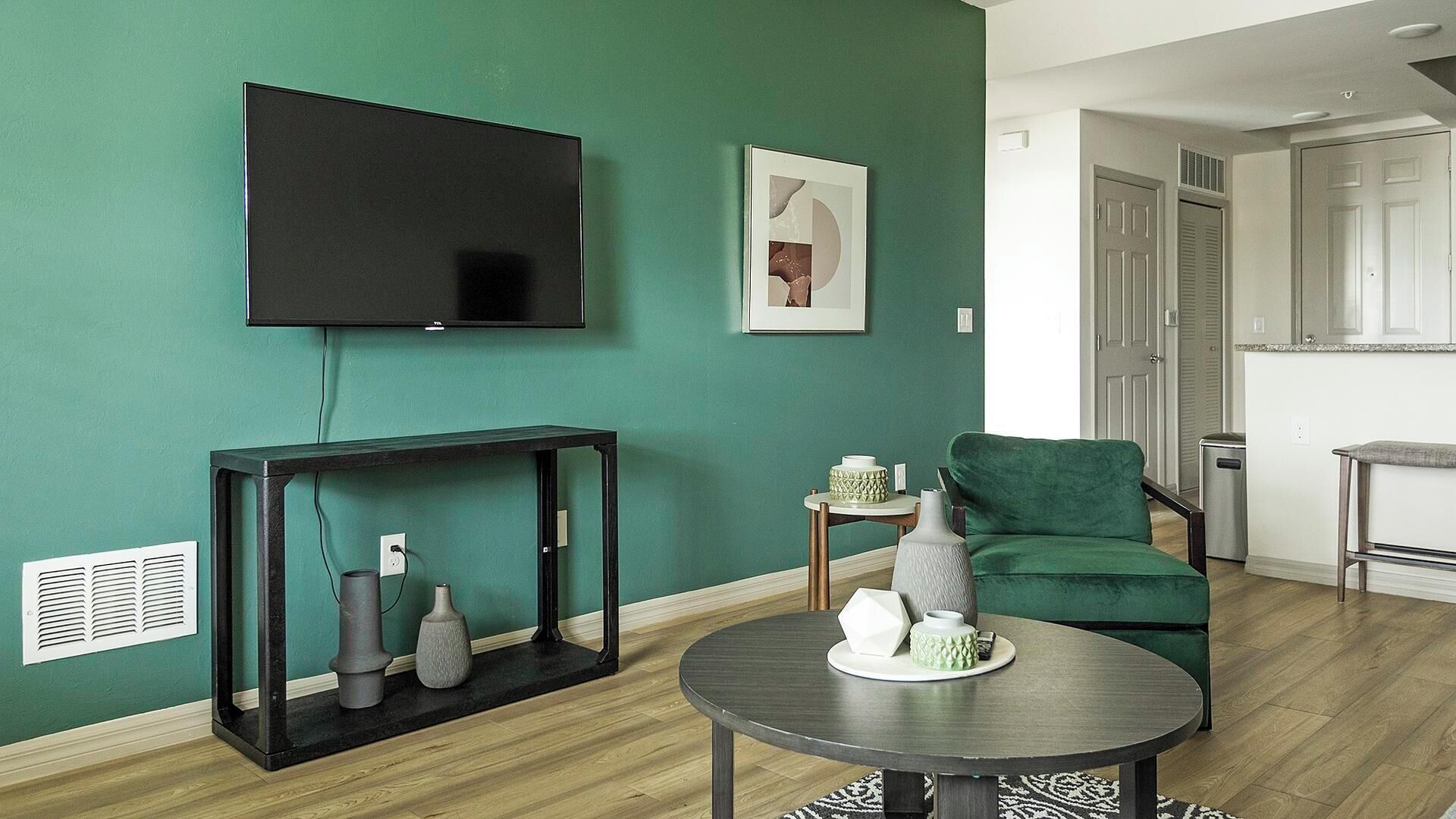 Un salon aux murs de couleur turquoise, doté d’un téléviseur à écran plat, d’une œuvre d’art encadrée, d’un canapé vert, d’une table basse agrémentée d’objets décoratifs et d’une console sur laquelle sont disposés des vases.