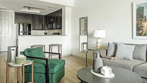 A modern living room with a green sofa, grey couch, and a round coffee table. There is a kitchen area with dark cabinets and a stainless steel refrigerator. A mirror and a lamp are on the wall.