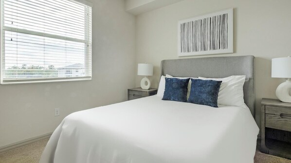 A neatly arranged bedroom with a large bed, two bedside tables, a window with blinds, and a wall-mounted artwork.