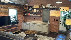 Una sala moderna con un sofá, una mesa de centro y una televisión. Se observa una zona de cocina con una pared de ladrillo, estanterías y una ventana.