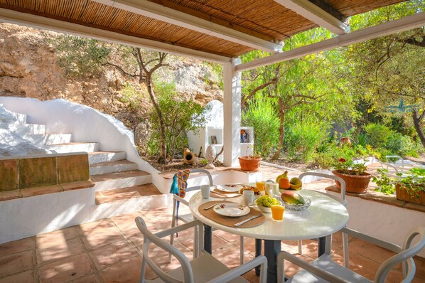 A covered outdoor dining area with a table set for two, surrounded by potted plants and a view of a rocky hillside.