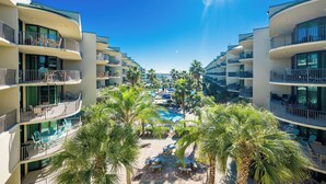Un resort con numerosi balconi, una piscina centrale circondata da palme e un cielo azzurro intenso.