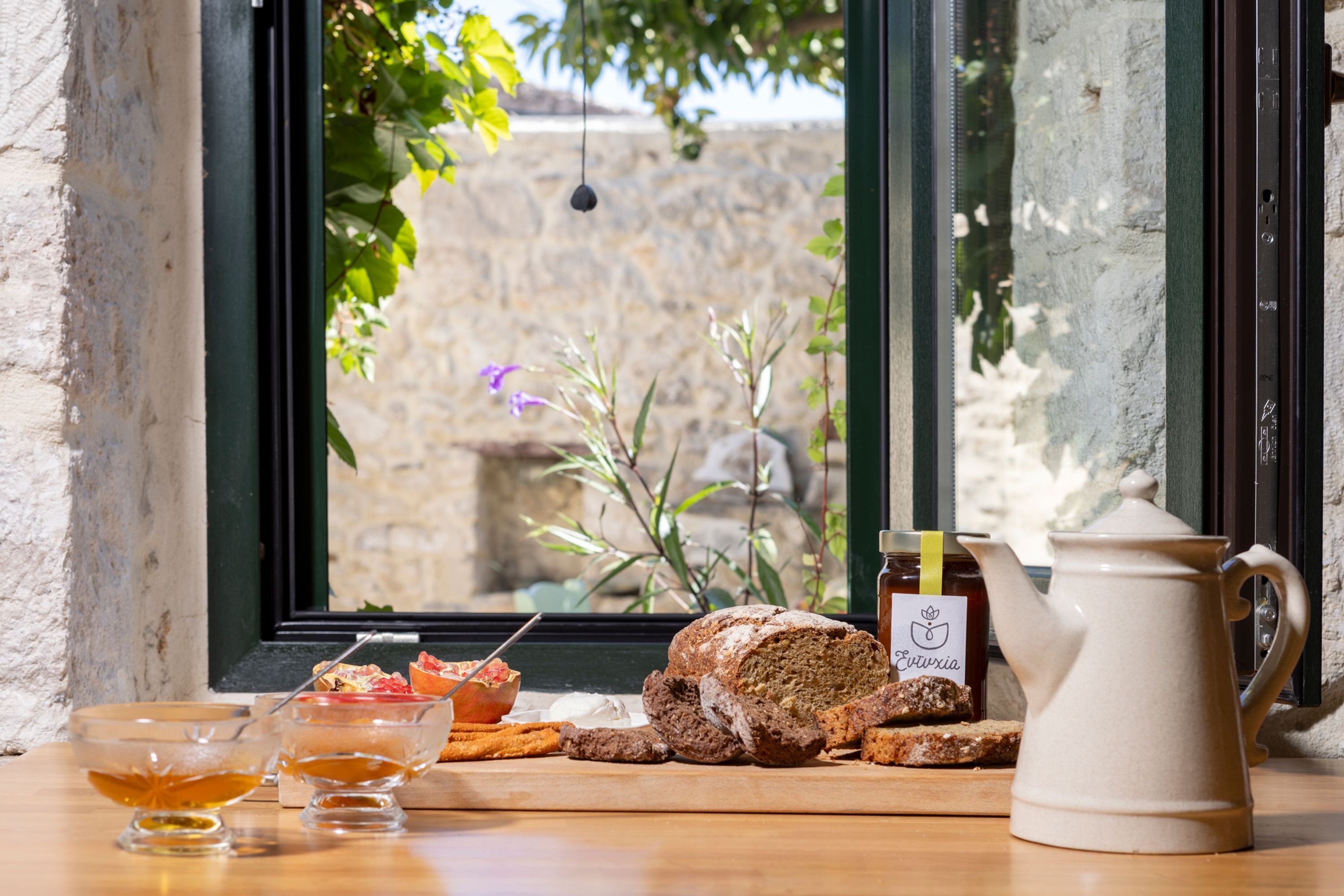A breakfast setting with bread, a teapot, and a jar of honey on a wooden table by a window with a view of a stone wall and greenery.