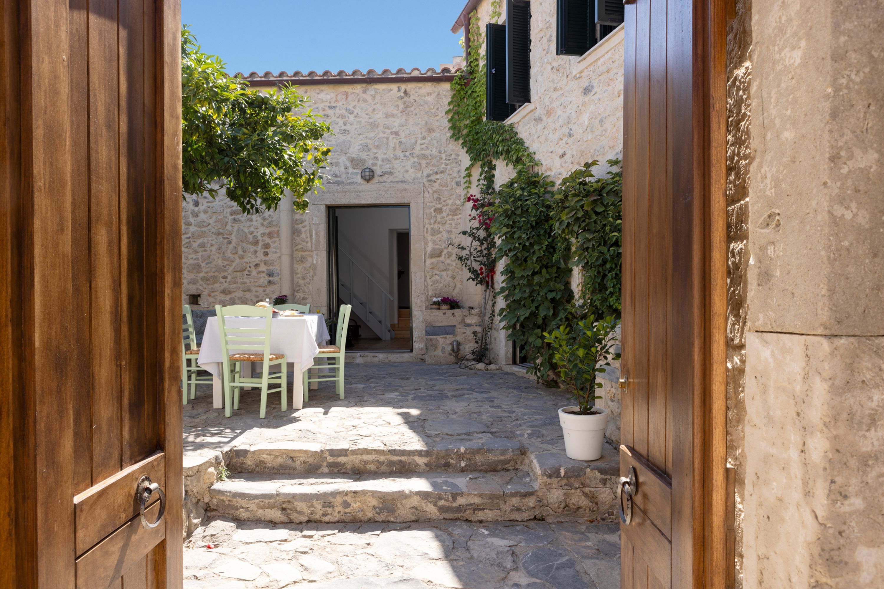 Une cour dallée avec une table et des chaises, un pot de fleurs et une porte donnant accès à un bâtiment.