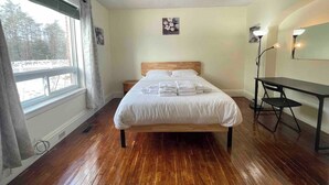 Un dormitorio con cama de madera, escritorio, silla, lámpara y ventana con vistas a árboles.